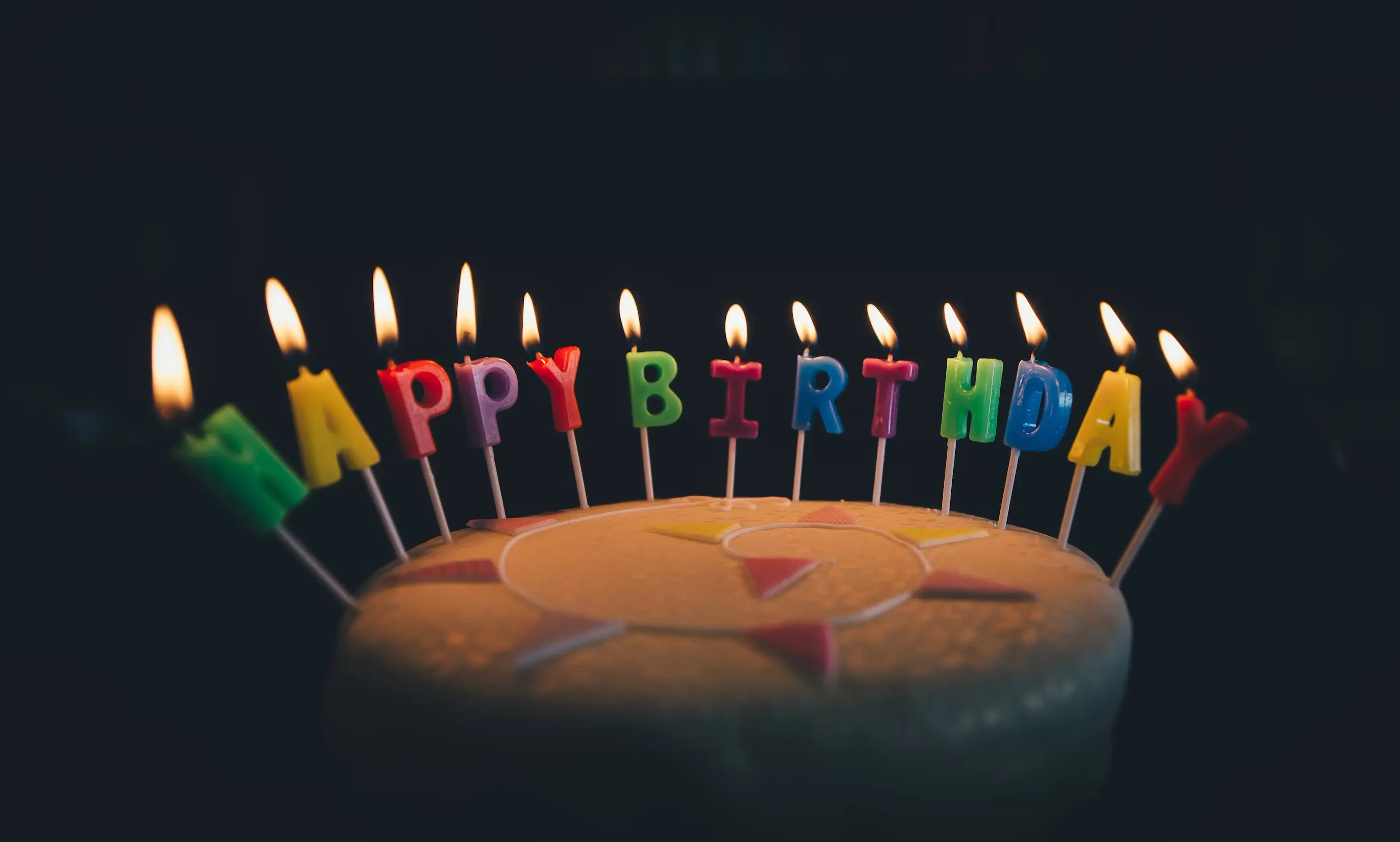 Birthday cake with colorful candles spelling out Happy Birthday, each candle lit against a dark background